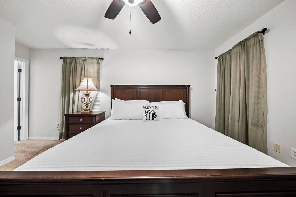 This cozy bedroom features a rich wooden bed frame complemented by soft white linens and a playful decorative pillow. Natural light filters through the warm-toned curtains, creating a tranquil ambiance perfect for restful nights.