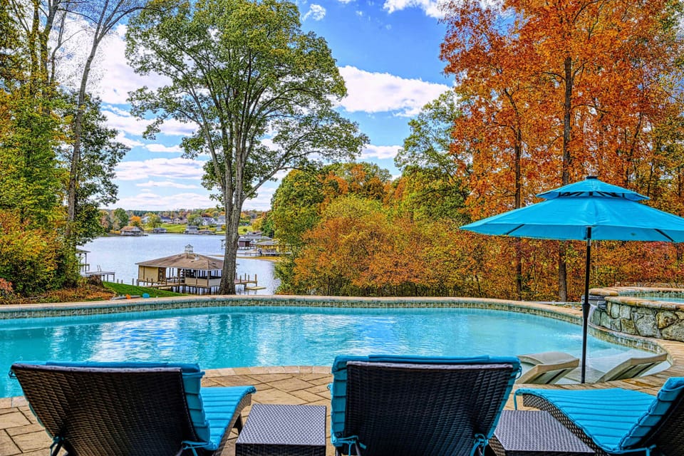 Resort-style heated pool with a built-in spa shelf and baja loungers in the water, framed by full lake views.