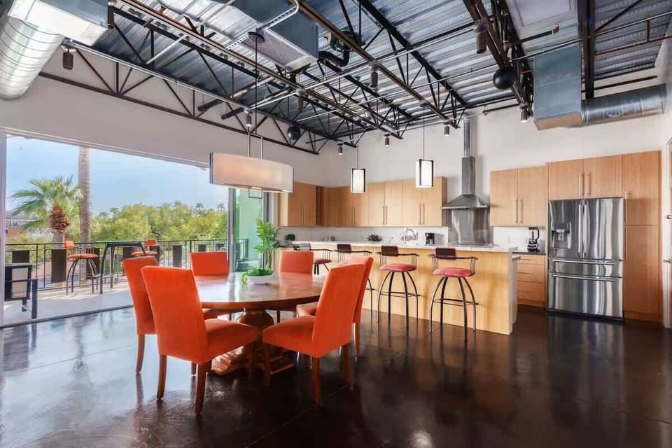 Dining area features a round table with seating for six in plush orange chairs, bathed in natural light with city views from the adjacent patio.