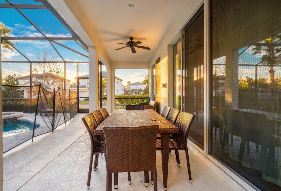 Enjoy a covered lanai with a wicker dining table, ceiling fan, and large sliding doors reflecting the pool area. Warm sunlight, outdoor seating, and peaceful views create a cozy spot for meals and relaxing Florida evenings.