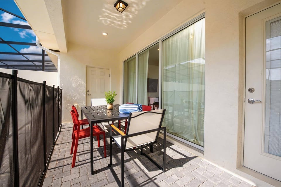 Enjoy outdoor meals on a covered patio with a black dining table, red chairs, sliding glass doors, pool view, and paved flooring, creating a relaxed space for vacation moments.