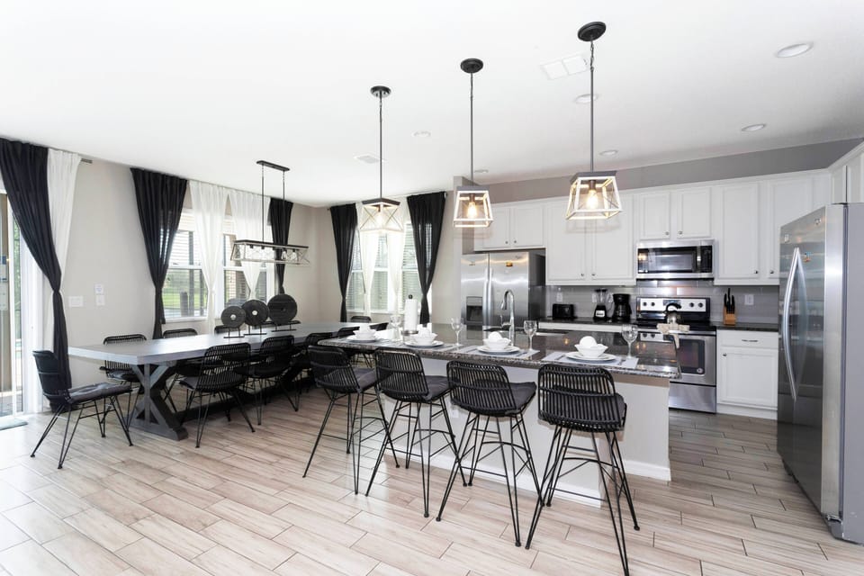This stylish kitchen boasts sleek stainless steel appliances and a spacious island with modern bar stools, ideal for casual meals and socializing, while large windows invite natural light, enhanced by elegant dark curtains.