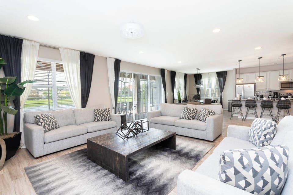 Experience a sunny living area with large windows that fill the space with natural light, showcasing soft gray sofas with stylish pillows. The modern coffee table and chic rug add a cozy, sophisticated touch, ideal for relaxation.