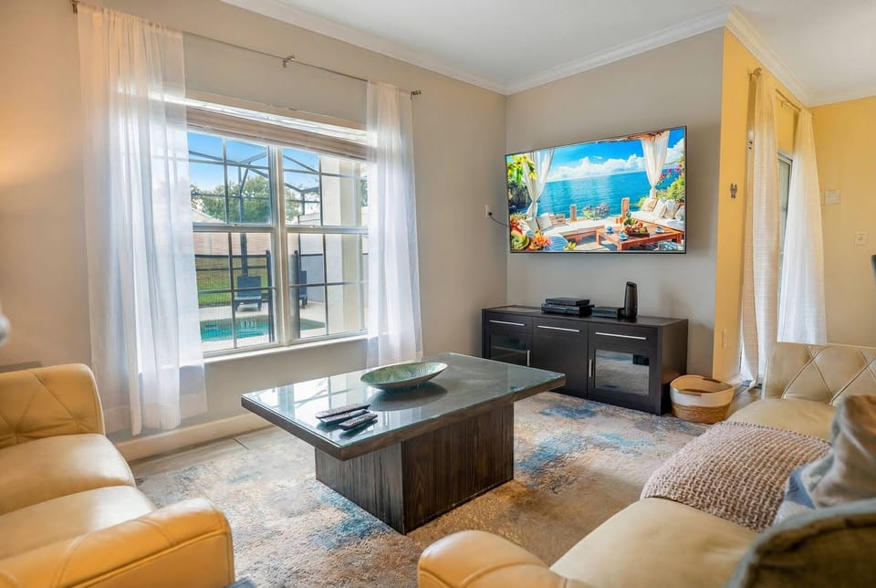 This cozy living room, bathed in natural light, features soft cream sofas and a stylish dark wood coffee table. A large flat-screen TV awaits your movie nights, while sheer curtains bring a touch of elegance to the space.