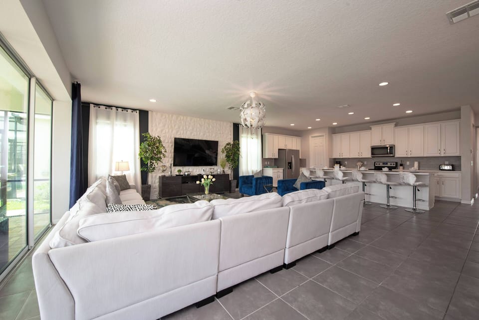 Relax in this bright living room featuring a plush white sectional and vibrant blue accent chairs, where large windows bring in natural light, creating a warm atmosphere perfect for gathering with friends and family.