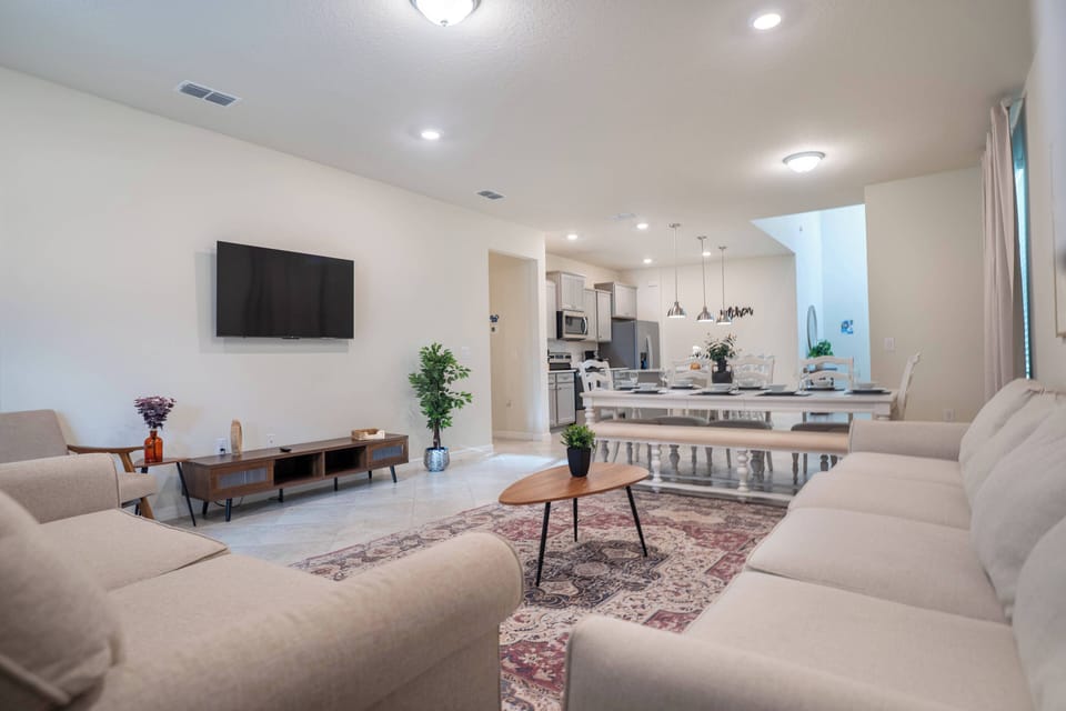 This bright and spacious living area boasts a soft beige sectional couch and a striking patterned rug, creating a cozy ambiance that invites relaxation and social gatherings, seamlessly flowing into the kitchen.