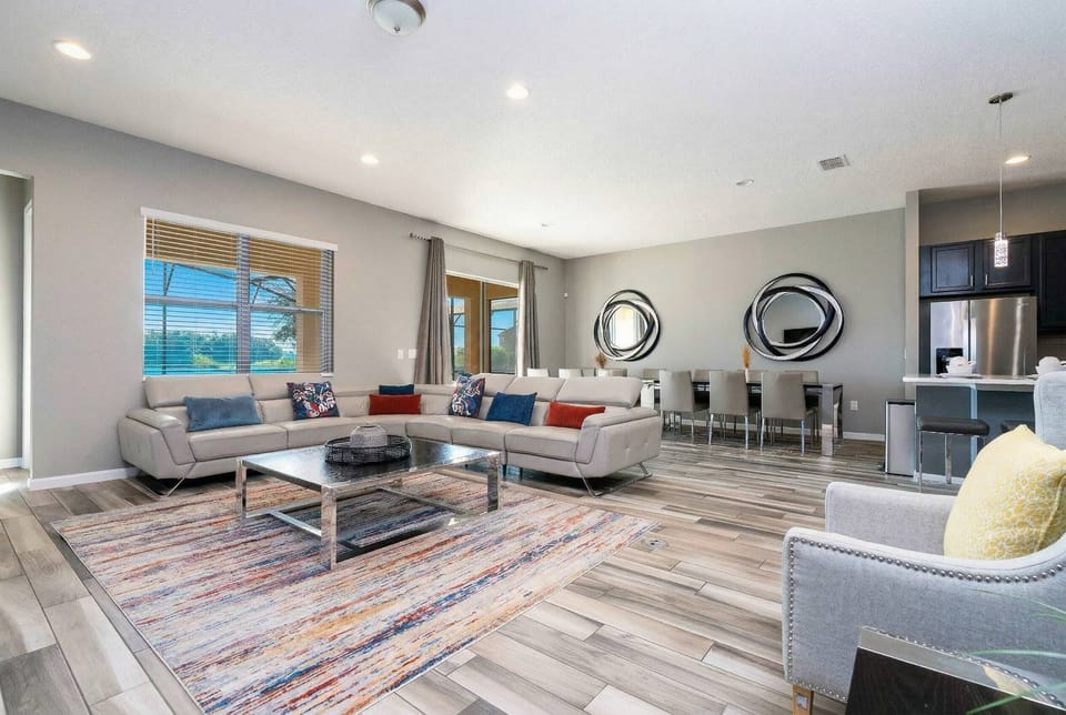 Sunlight fills this spacious living area, showcasing soft gray sofas with colorful pillows and a chic rug. With inviting mirrors and a smooth transition to the dining area, it’s an ideal spot for relaxation and socializing.