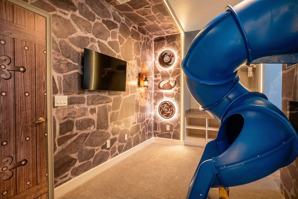 Welcome to a whimsical playroom with a vibrant blue slide and a charming stone-patterned wall. The cozy carpet and unique decor make it the perfect space for family fun and relaxation together. Enjoy your time!