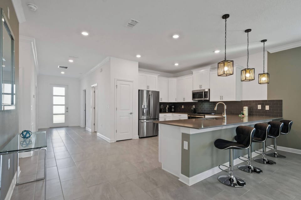The spacious kitchen boasts sleek white cabinetry and a dark countertop, offering a stylish contrast. Enjoy casual meals at the modern bar with chic black stools, all warmly lit by elegant pendant lights for a cozy ambiance.