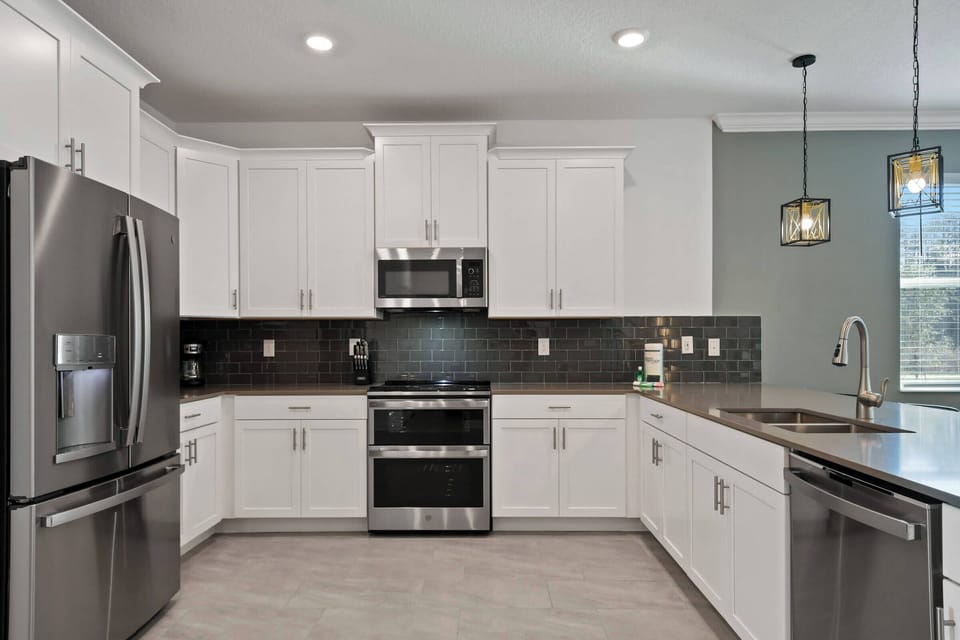 Enjoy a stylish kitchen with sleek stainless steel appliances and elegant white cabinetry, accented by a striking dark backsplash. The subtle pendant lighting adds a modern touch, creating a welcoming space for meal preparation.