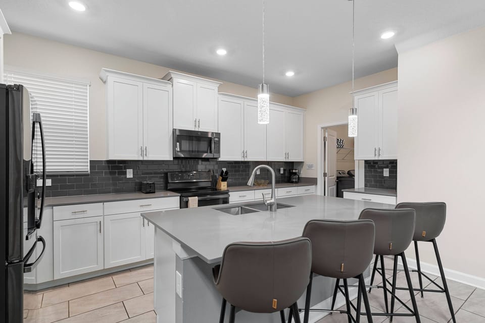 The kitchen boasts a spacious island with sleek gray countertops and stylish bar stools, ideal for casual dining. Bright white cabinetry beautifully contrasts with dark appliances, creating a fresh and functional cooking space.