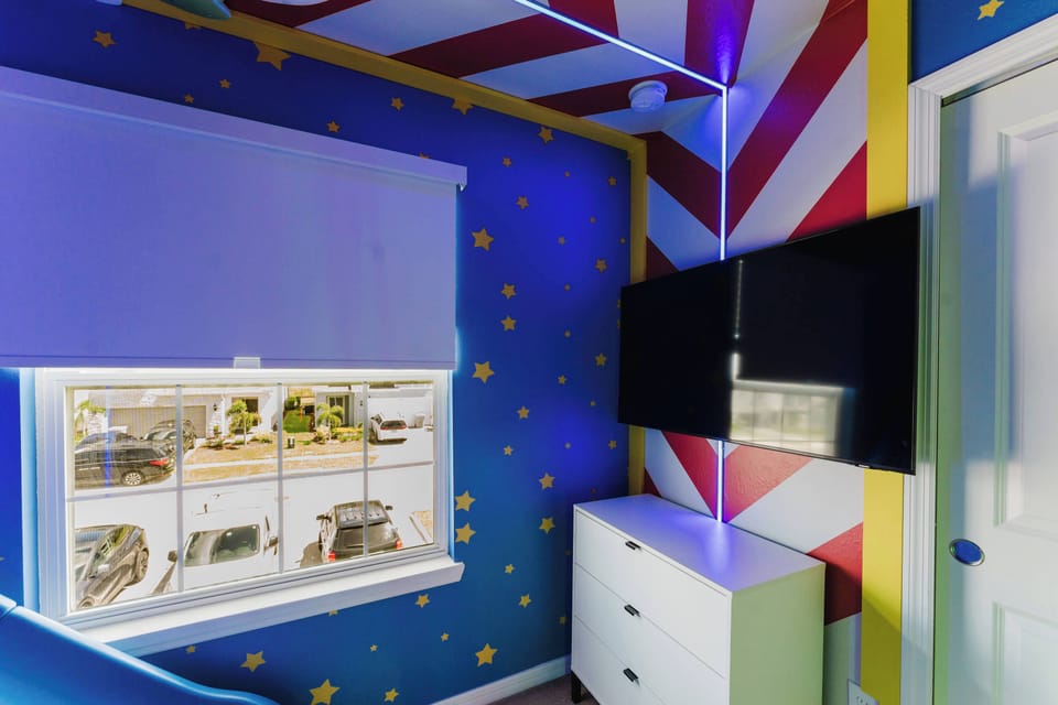Enjoy a vibrant room with cheerful blue walls and golden stars that uplift your spirit. The sleek white dresser contrasts with a striking red and white striped ceiling, while a large window invites the outdoors in, adding to its.