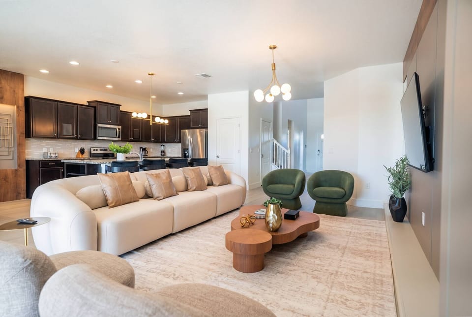 Relax in a bright open living area with a large sofa, green accent chairs, wall-mounted TV, modern lighting, and a nearby kitchen, creating a comfortable space to gather during your vacation.
