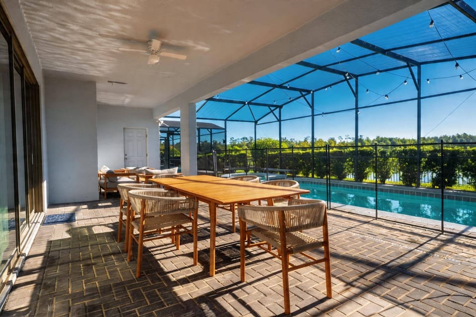 Dine outdoors on a covered patio with a large table, comfortable seating, ceiling fan, screened pool views, string lights, and peaceful greenery, creating a perfect space for vacation meals.