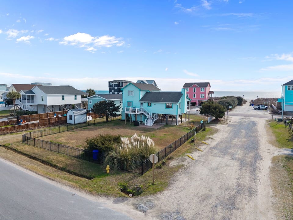 Aerial view of a turquoise raised cottage with deck stairs, large fenced lawn, gravel road to beach access, neighboring coastal homes, and ocean views beneath a bright blue sky.