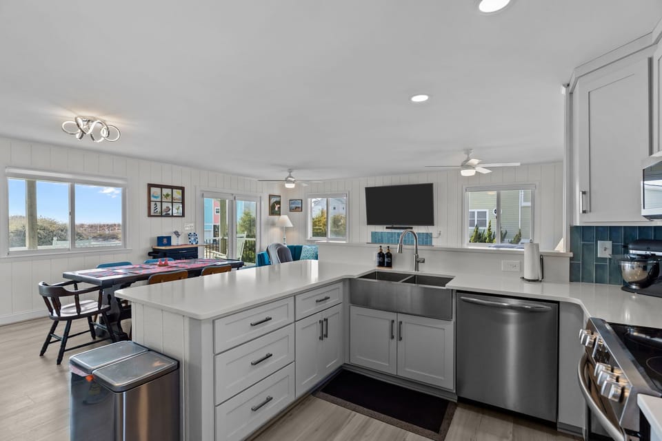 Bright beach house kitchen with white counters, gray cabinets, blue tile backsplash, stainless appliances, double sink, window views, and turquoise bar stools beside the open living area.