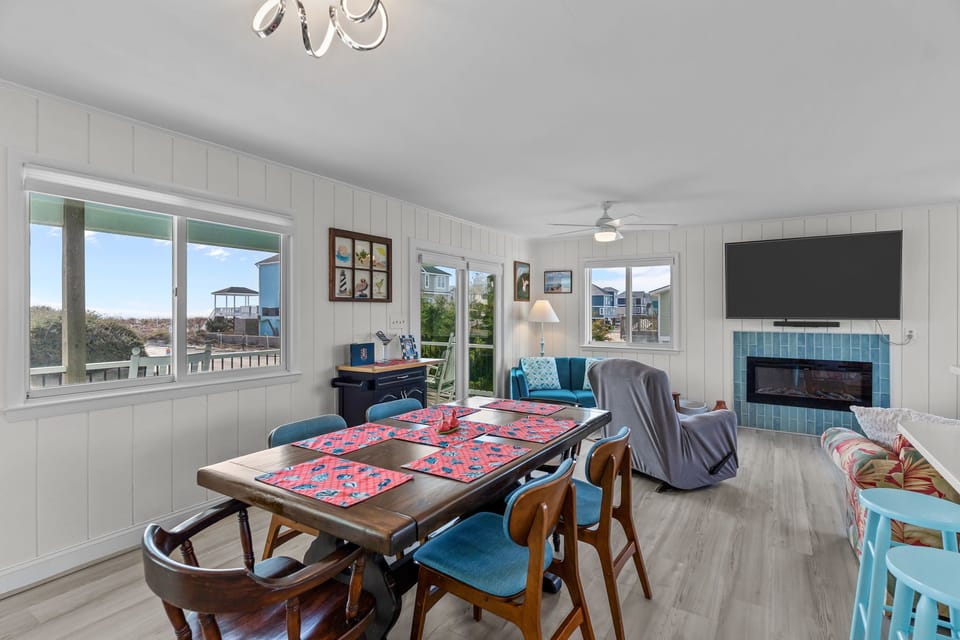 Bright open interior with wood dining table, blue chairs, ocean-view windows, sliding door, turquoise loveseat, armchair, wall-mounted TV, blue-tile fireplace, bar stools, and light wood floors.