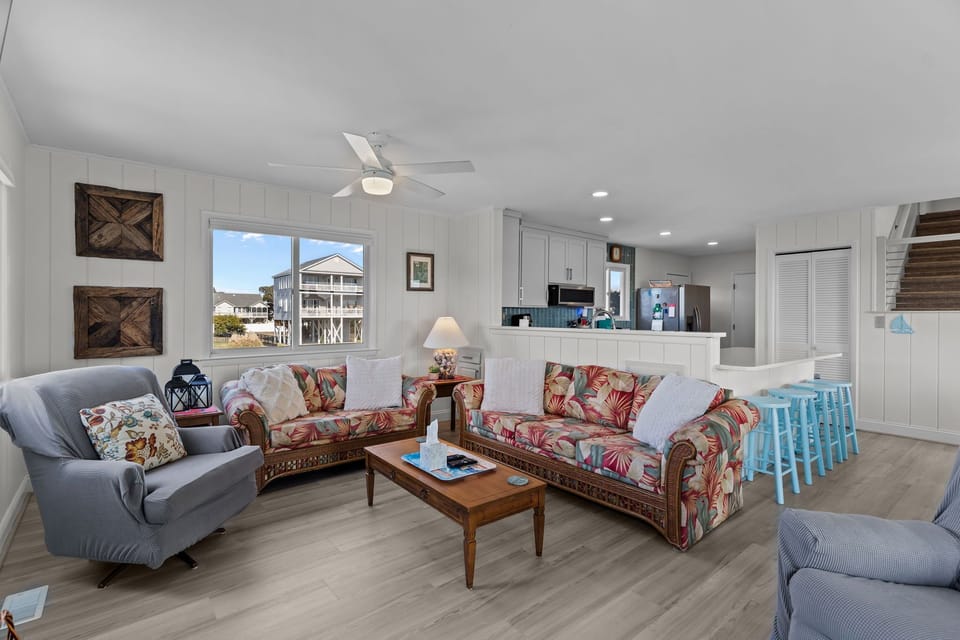 Bright open interior with wood dining table, blue chairs, ocean-view windows, sliding door, turquoise loveseat, armchair, wall-mounted TV, blue-tile fireplace, bar stools, and light wood floors.