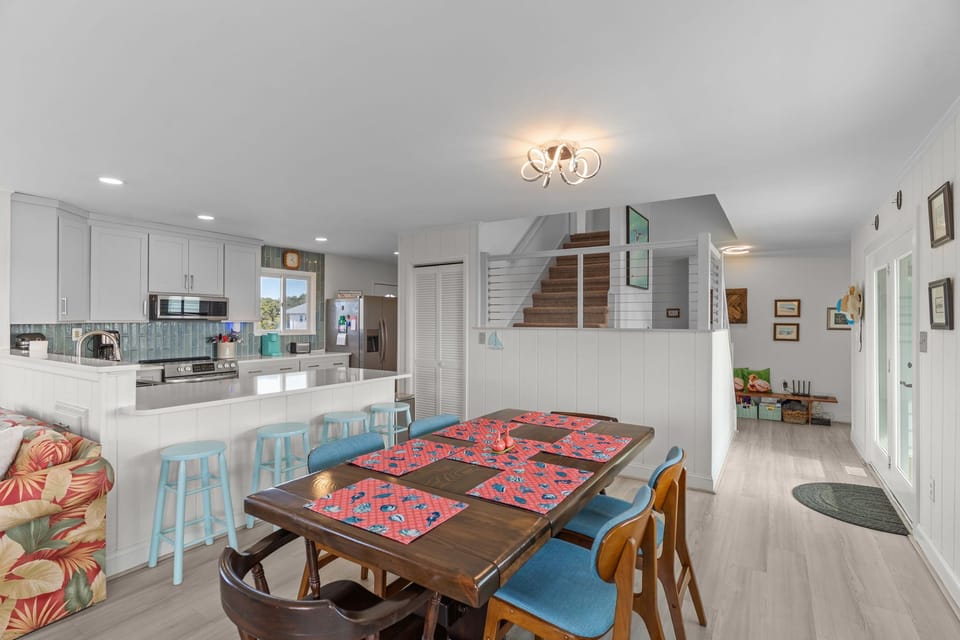 Bright open interior with wood dining table, blue chairs, ocean-view windows, sliding door, turquoise loveseat, armchair, wall-mounted TV, blue-tile fireplace, bar stools, and light wood floors.