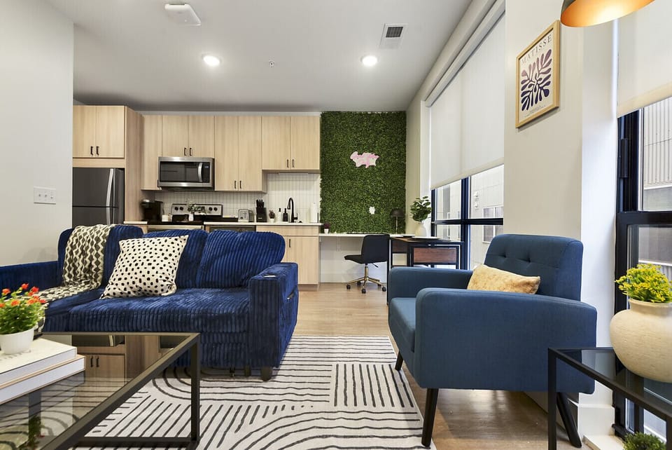 Cozy downtown living area with deep navy corduroy sectional sofa, chic glass coffee table, vibrant green plant wall accent, and open flow to a modern workspace—perfect for relaxing or remote work.