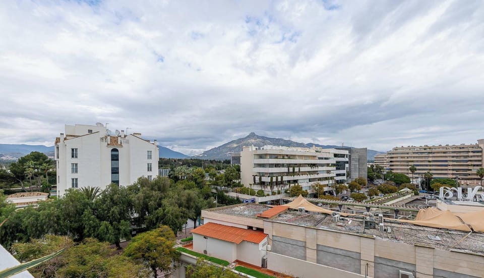 Urban view with mid-rise buildings, greenery, and distant mountains under cloudy skies. A balanced mix of city and nature creates a modern, scenic backdrop.