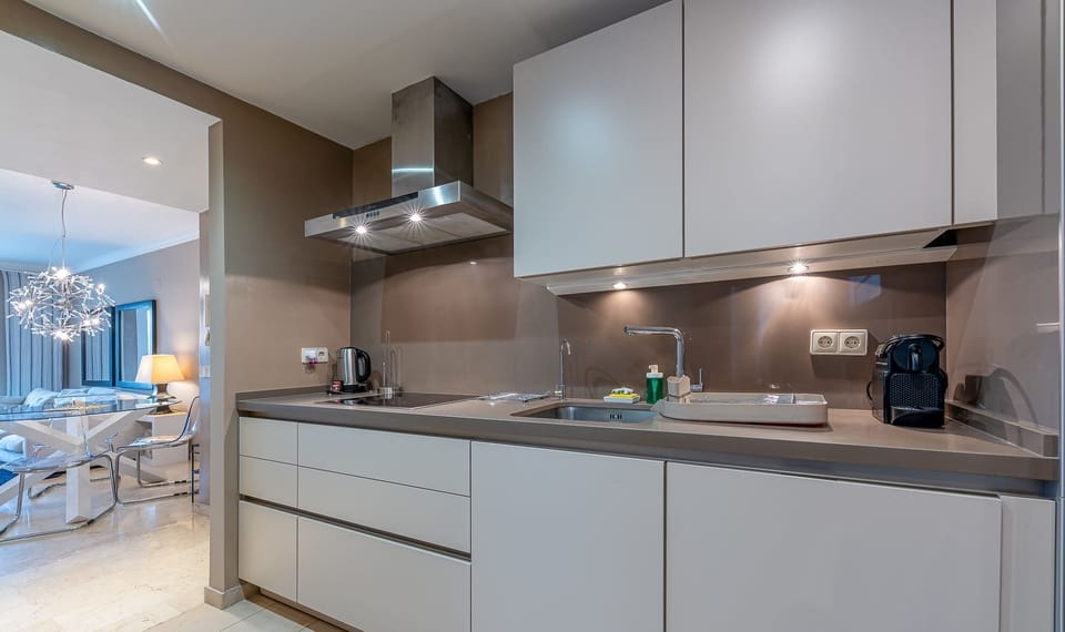 Modern kitchen with white cabinetry, gray counters, stainless range hood, and taupe backsplash. Warm lighting and sleek design create a bright, inviting cooking space.