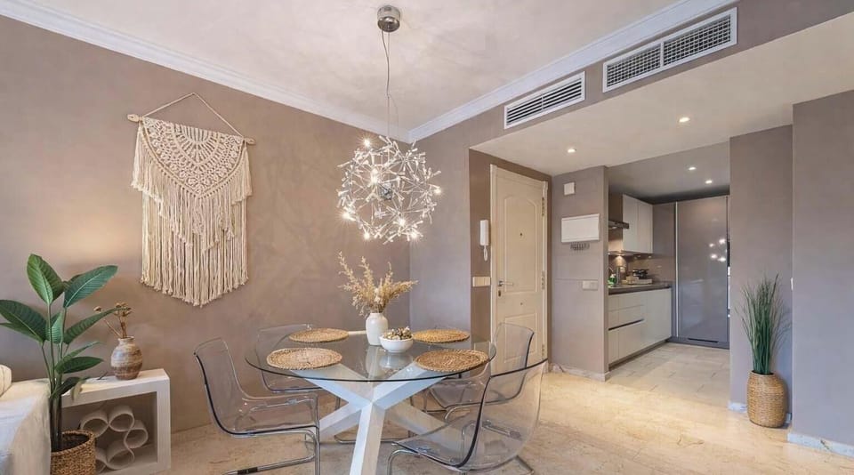 Stylish dining-kitchen space with round glass table, transparent chairs, macramé wall art, and modern chandelier. Natural accents and sleek cabinetry create a bright, inviting design.