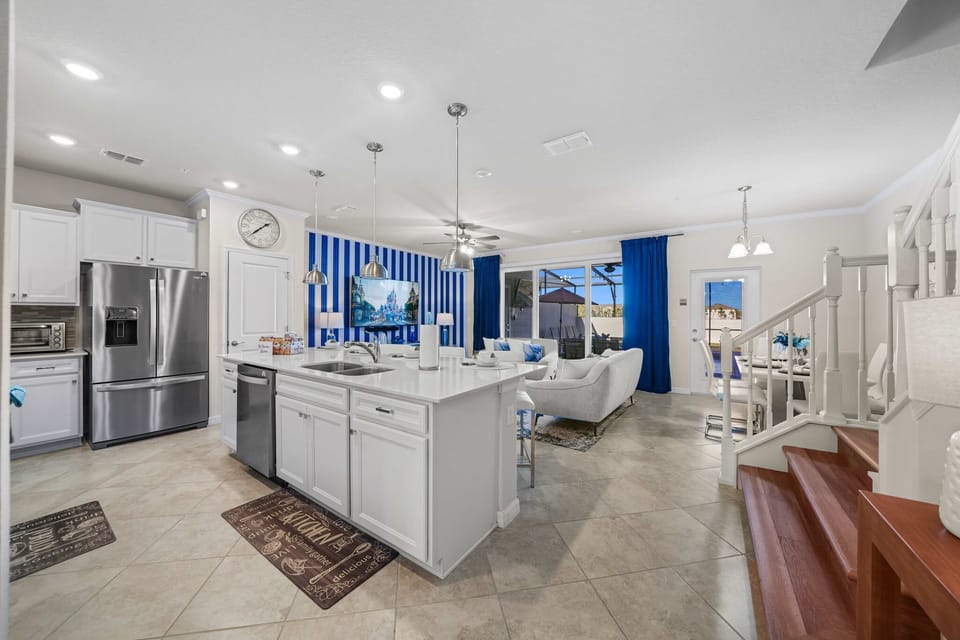 Bright open-concept hub: sleek white kitchen with stainless appliances and island seating, flowing to a comfy lounge with bold blue accents and patio access.
