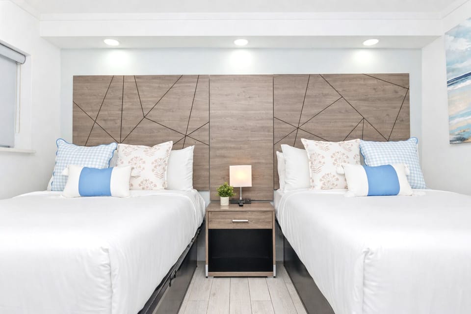Bright, modern bedroom with two cozy queen beds & a sleeper sofa, a stylish wood accent wall, soft linens, bedside lighting, and a blackout shade—perfect for a restful stay.