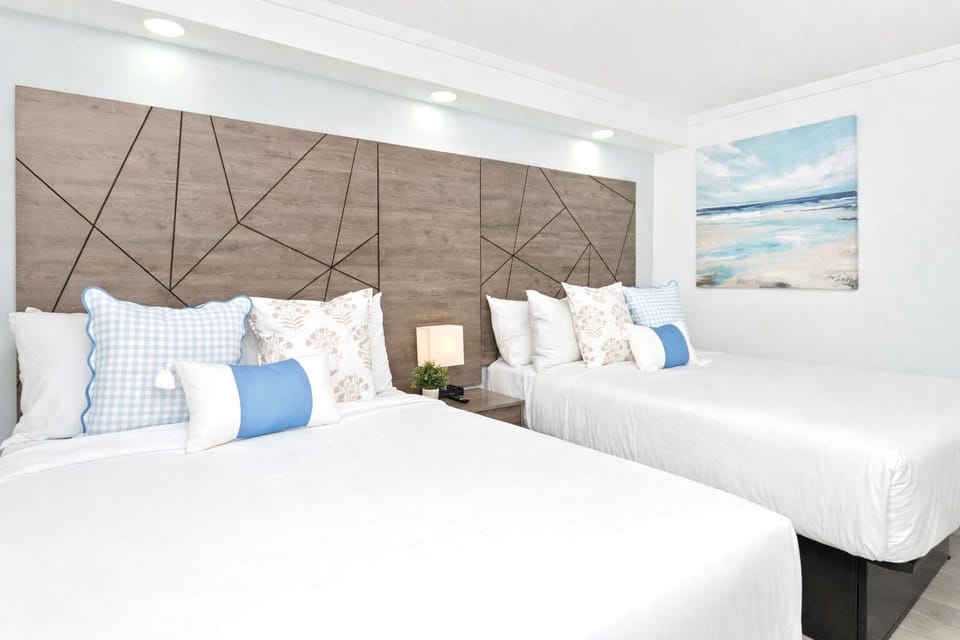 Bright, modern bedroom with two cozy queen beds & a sleeper sofa, a stylish wood accent wall, soft linens, bedside lighting, and a blackout shade—perfect for a restful stay.