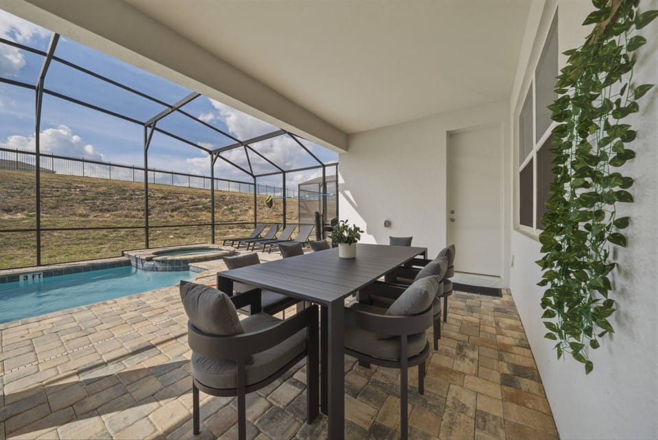 Experience outdoor dining in a stylish setting with a sleek black table and comfy chairs. Sunlight streams through a protective screen, brightening the inviting pool area and lush greenery, creating the ideal spot for relaxation.