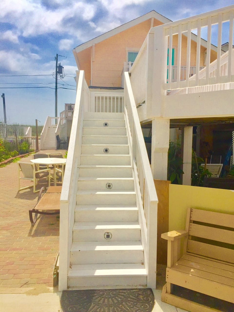 Modern beach house exterior with white stairs leading to upper deck and outdoor seating areas in sunny coastal setting.