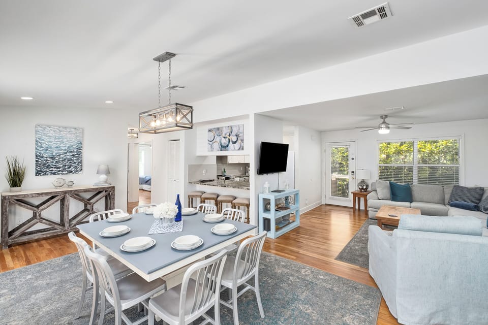 Unwind in this bright, open living space where warm wood floors meet coastal-inspired decor. Gather around the dining table for meals, relax on the comfortable sofa, and enjoy sunlight streaming through large windows.