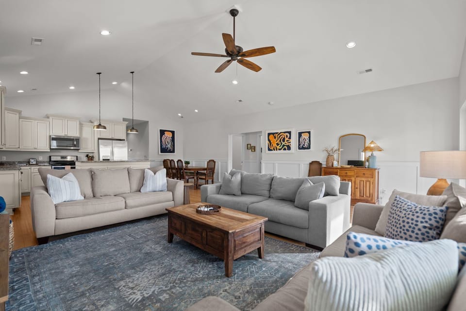 Bright open-concept living space with vaulted ceilings, cozy seating, and coastal-inspired décor. The living room flows seamlessly into the kitchen and dining area, creating an inviting space for relaxing and gathering.