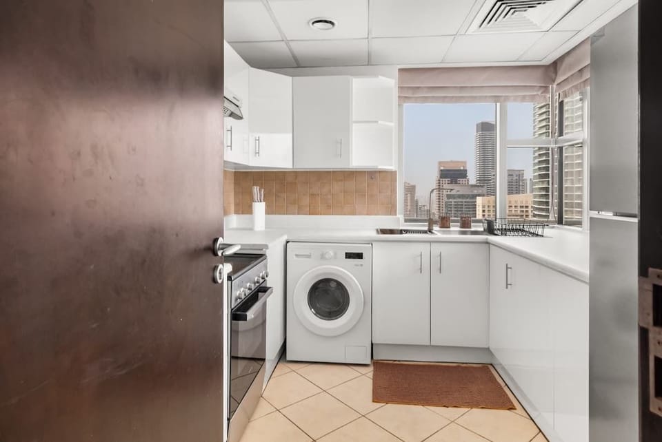  A compact modern kitchen with sleek white cabinets, tiled backsplash, and essential appliances including a stove, washing machine, and fridge. Large windows brighten the space, offering city views and a fresh, airy atmosphere.