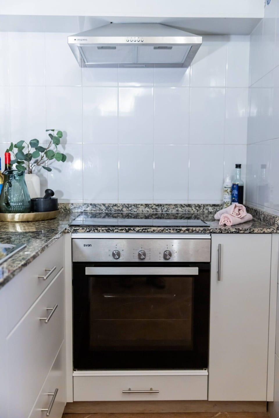Küche OG | Nahaufnahme Kochbereich: Svan-Backofen (stainless, Drehregler) + Cerankochfeld + Oopes-Dunstabzug (stainless) + weiße Fliesenrückwand + Granit-Theke mit Eukalyptus + grüner Vase