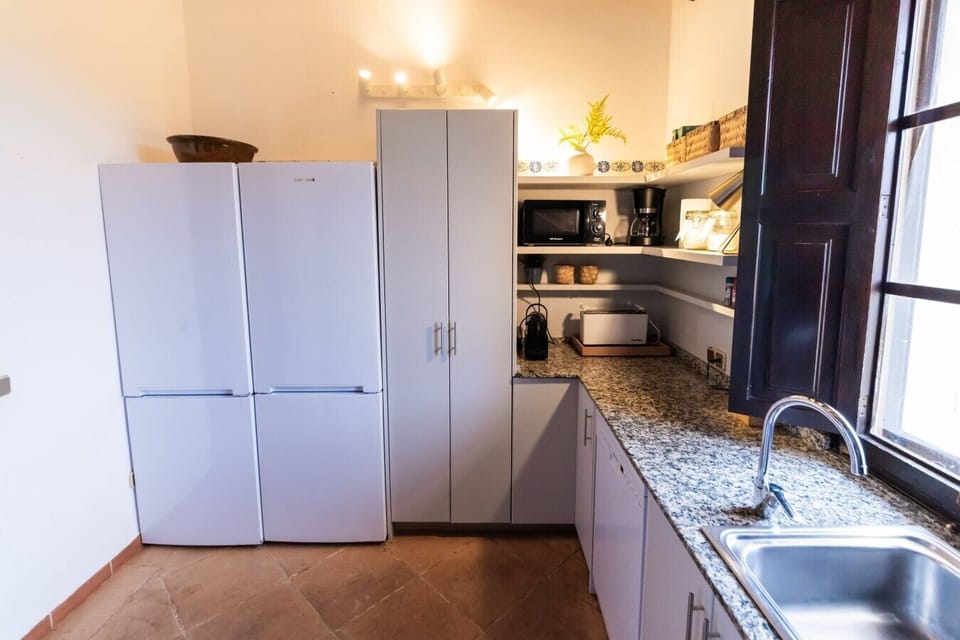 Küche OG | 2 weiße Kühlschränke nebeneinander + hoher weißer Vorratsschrank, Gerätecke (Mikrowelle schwarz + Kaffeemaschine + Toaster) auf Regalen + Granit-Arbeitsplatte + Edelstahl-Spüle rechts + Terrakottaboden