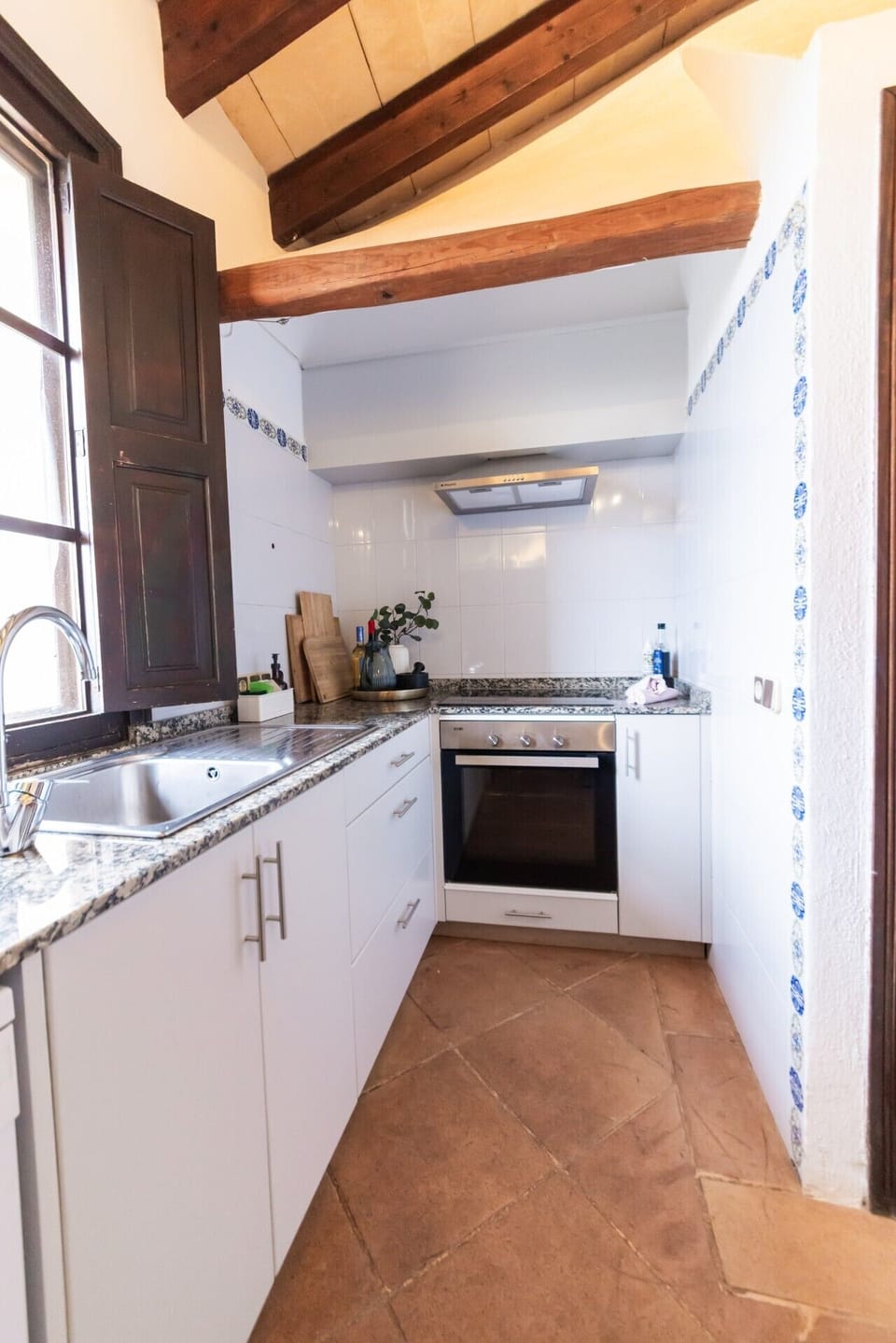 Küche OG | Weitwinkel Kochbereich: Edelstahl-Spülbecken links + Svan-Backofen + Dunstabzug + weiße Fliesen + blaue Azulejo-Leiste + dunkle Holzläden links, Holzbalken-Decke + Steinboden