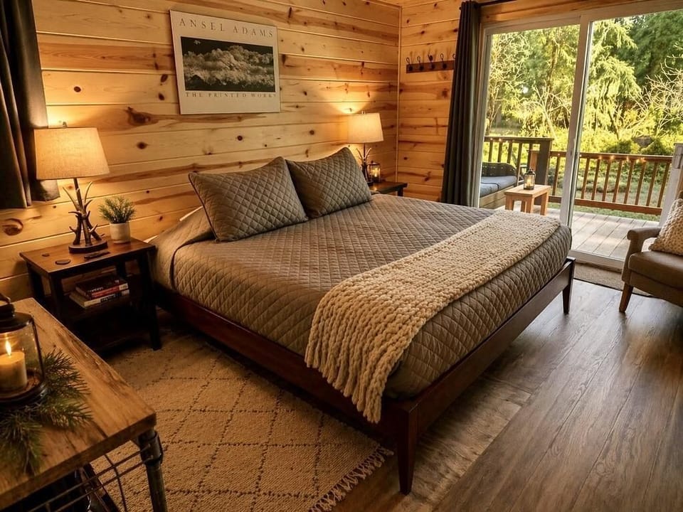 King bed with quilted gray bedding sits beside wood-paneled walls and sliding glass doors to a deck with forest views.