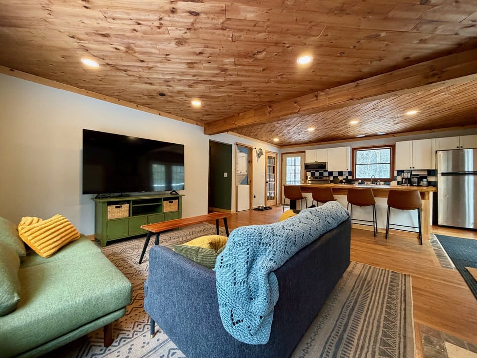 The open-concept living and kitchen area features wood tones, comfortable furniture, a large Smart TV set, and an island with bar stools.