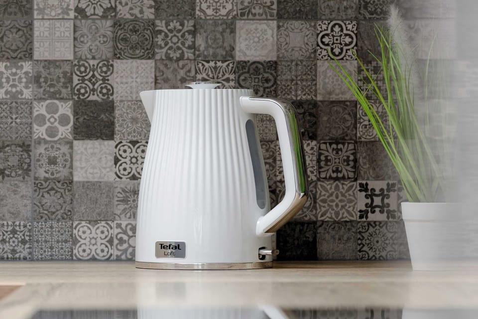 A close-up of an electric kettle placed on a kitchen counter. Patterned tiles form a decorative backdrop. The scene emphasizes everyday convenience.