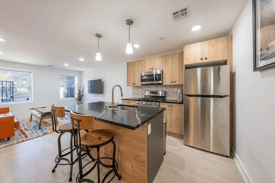 The kitchen features a dark stone island with two barstools and minimalist pendant lighting. Stainless steel appliances and light wood cabinetry provide a clean, functional space for meals.