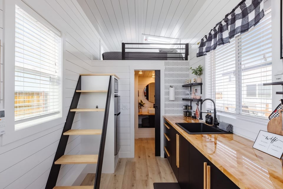 Clean and modern kitchen with loft access, butcher block countertops, black cabinetry, and oversized windows for natural light. Open-concept tiny home design creates a bright, airy feel perfect for couples and weekend escapes.