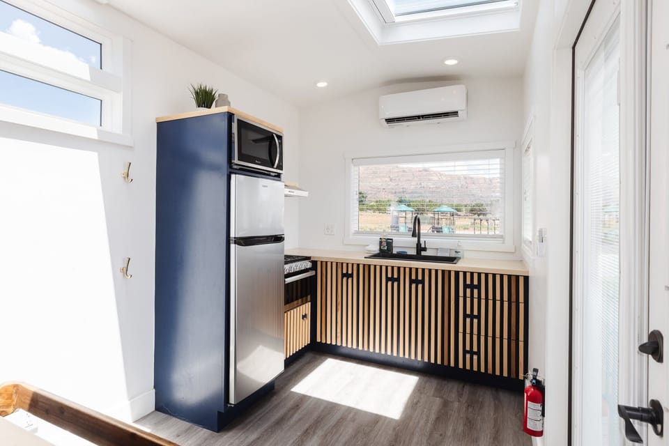 Bright and modern tiny home kitchen featuring a refrigerator, stove, oven, microwave, sink, and ample counter space. Large windows fill the space with natural light, creating a welcoming area for meal preparation during your Southern Utah getaway.