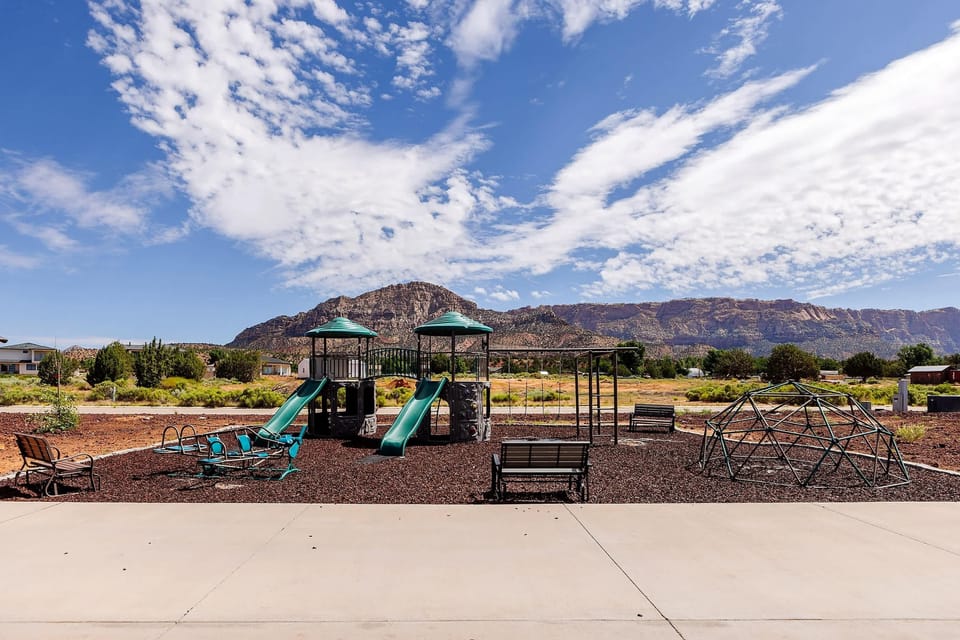 Family-friendly playground with slides, swings, a climbing dome, and an airplane teeter-totter. Benches nearby provide a comfortable place for parents to sit while kids enjoy outdoor play just steps from the tiny homes.