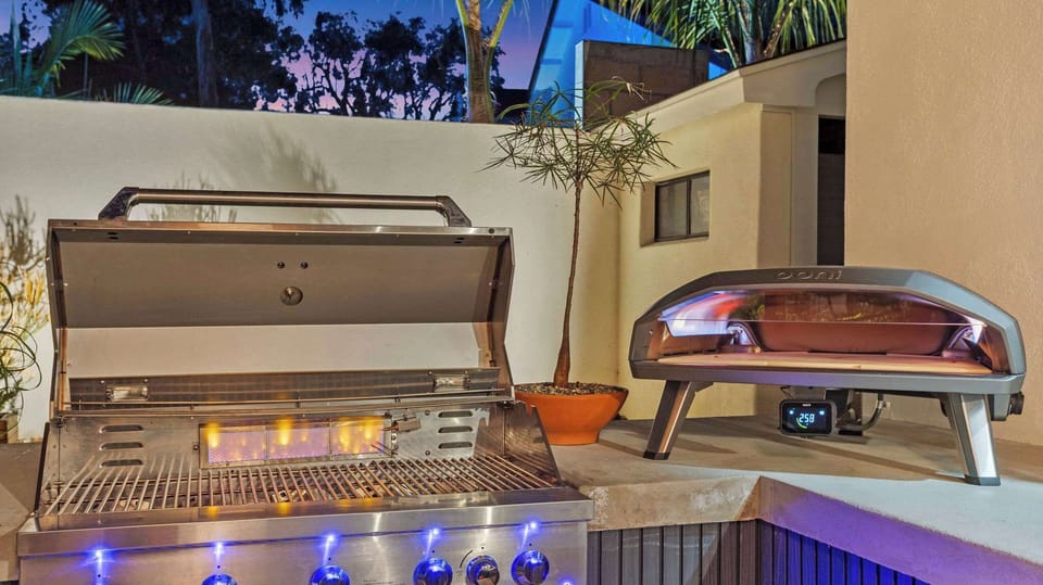 Outdoor kitchen at dusk — stainless gas grill and lit Ooni pizza oven fired up and ready. Group dinners just got an upgrade