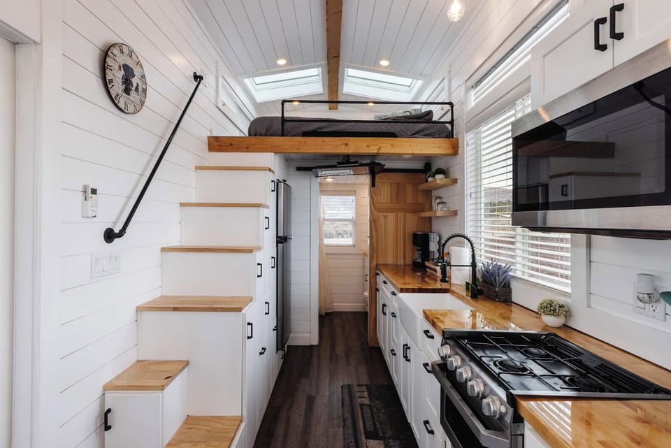 Spacious tiny home kitchen featuring a full gas range, stainless appliances, open shelving, and smart storage beneath the staircase for a functional, comfortable stay.