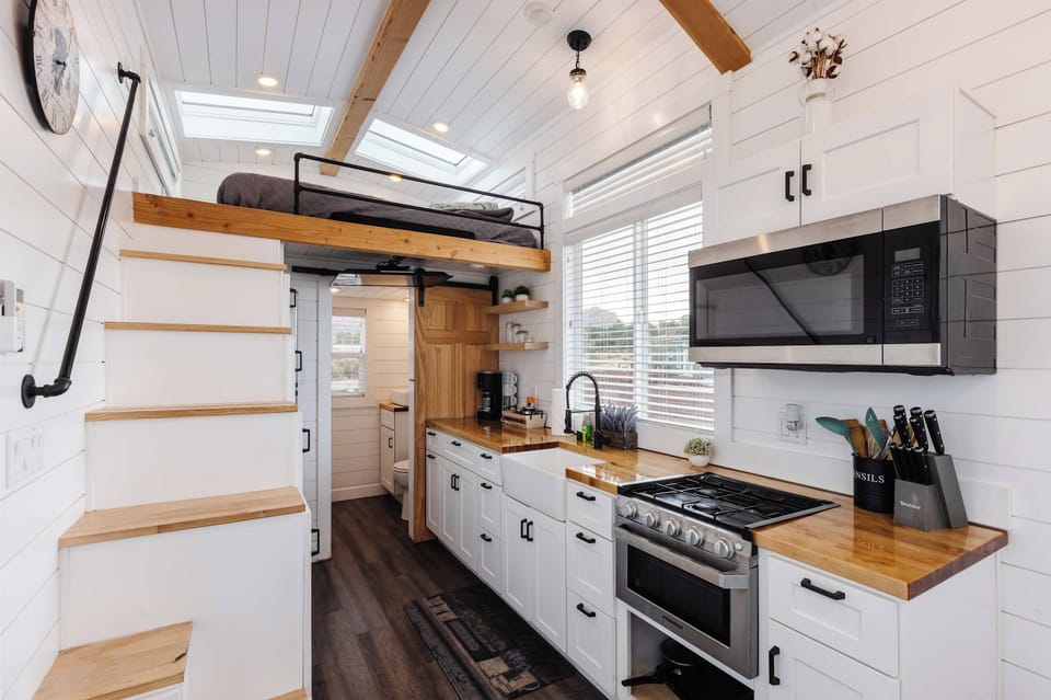 Bright full kitchen with gas range, microwave, farmhouse sink, and butcher block countertops. Stairs lead to the loft bedroom, maximizing space in this modern tiny home near Zion.