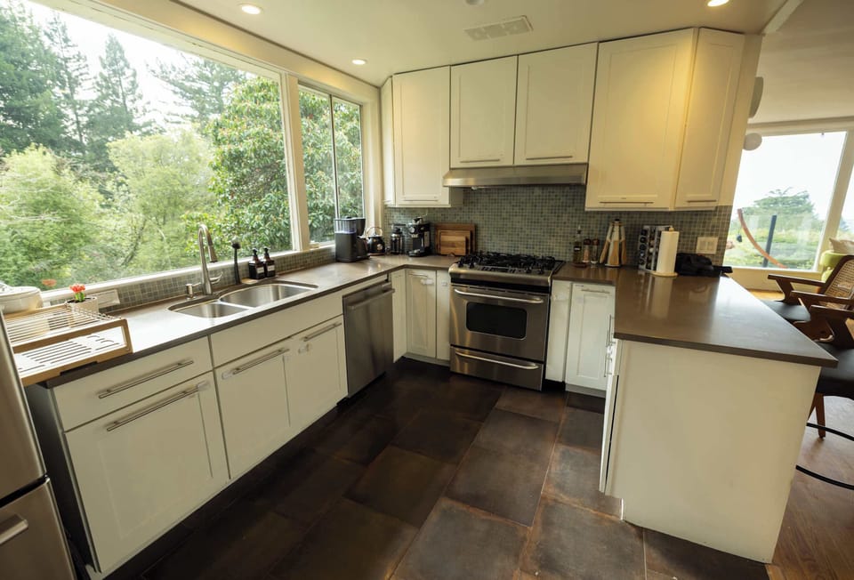 This bright kitchen features expansive windows with lush views, sleek cabinetry, modern appliances, and generous counter space, making it a welcoming spot to cook and gather
