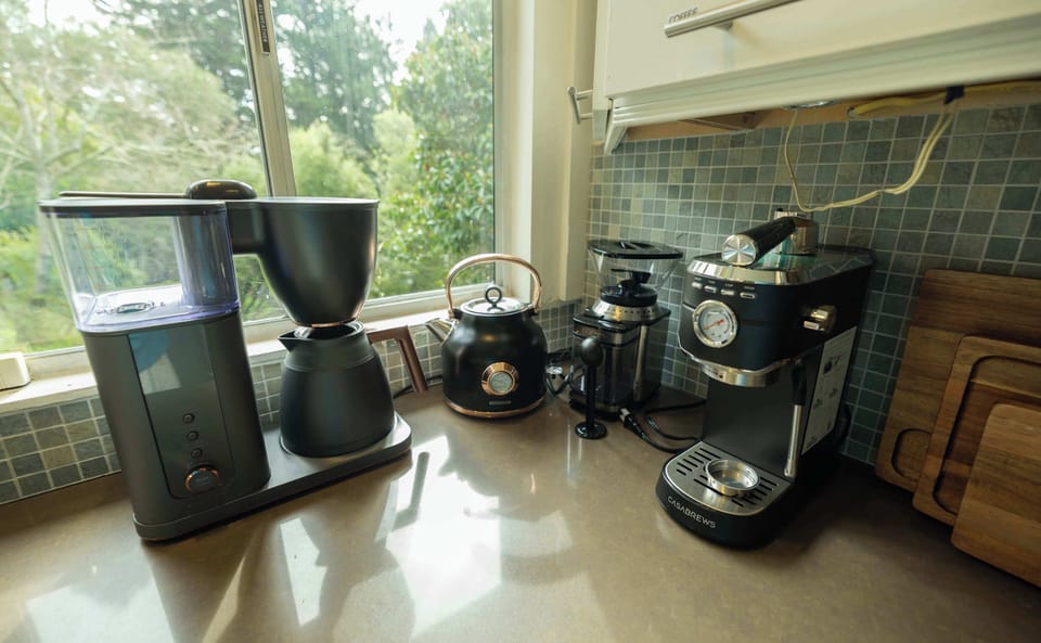A fully equipped coffee station with a drip maker, kettle, and espresso machine sits beside sunlit windows—perfect for starting your morning with your favorite brew and a peaceful view